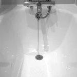 A close-up of a white bathtub with a metal chain attached to the drain plug, faucet, and handheld showerhead above.