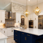 Modern kitchen with white cabinets, blue island, gold fixtures, stainless steel appliances, patterned backsplash, and two pendant lights over the island.