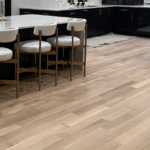 Modern kitchen with light wood flooring, a large marble island with four white barstools, dark lower cabinets, and a marble backsplash.