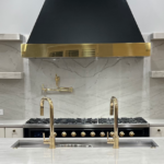 Modern kitchen with marble countertops and backsplash, gold and black range hood, double sink, gas stove, and arched doorways.